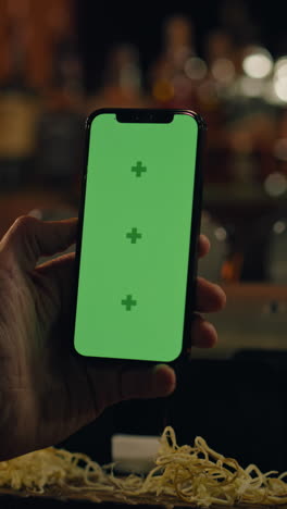 Vertical-shot-of-man-holding-phone-with-green-screen,-touching-display.-Sports-fan-watches-bookmaker-ratings-online-or-surfs-the-Internet,-talks-with-bartender-sitting-at-bar-counter-in-pub.-Chroma-key.