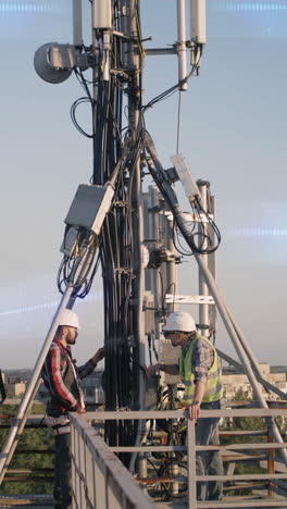 Tilt-up-view-of-men-in-hardhats-repairing-modern-antenna-transmitting-data-against-sunset-sky.-Vertical-shot