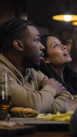 Vertical-shot-of-friends-rejoice-while-watching-live-sports-match-on-TV-in-bar-or-pub-in-the-evening.-Happy-sports-fans-applaud-and-shout-when-football-team-scores-goal-in-world-league-tournament.