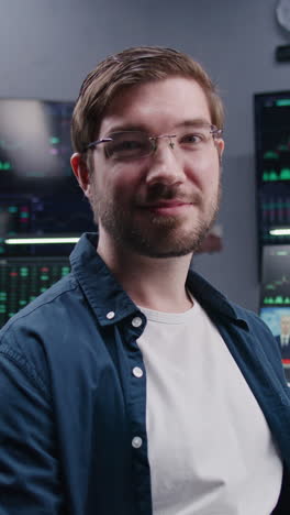 Male-financial-analyst-sits-at-workplace,-talks-into-the-camera-and-look-at-it.-Coworkers-analyze-exchange-market-charts-on-big-screens-at-background.-Vertical-shot.-Portrait.