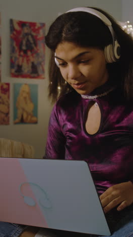 Beautiful-African-American-girl-in-headphones-sits-on-bed-in-her-room-and-surfs-internet-using-tablet.-Happy-teenager-chats-with-friends-or-listens-music.-Vertical-shot