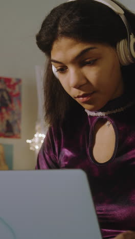 Vertical-video-of-teenage-girl-in-headphones-chatting-with-friend-online-and-surfing-the-Internet-on-laptop-sitting-on-the-bed-in-bedroom.-African-American-girl-spends-leisure-time-having-fun-at-home.