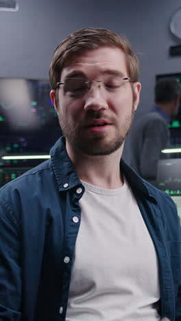 Male-financial-analyst-sits-at-workplace,-gesticulate,-talks-into-the-camera-and-look-at-it.-Coworkers-analyze-exchange-market-charts-on-big-screens-at-background.-Vertical-shot.-Portrait.