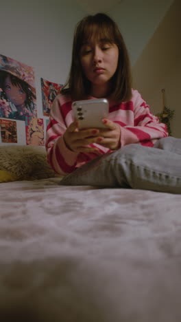 Asian-girl-uses-smartphone-chatting-or-browsing-social-media,-then-locks-phone-and-rests-in-bed-in-bedroom.-Teenage-girl-spending-leisure-time-at-home-after-school.-Lifestyle-concept.-Vertical-video.
