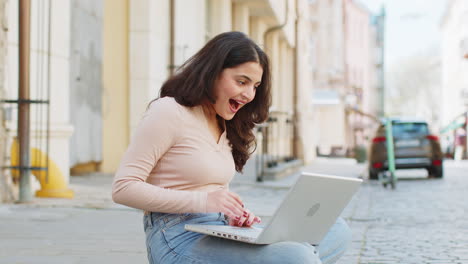 Glückliche-Indische-Frau,-Die-Am-Laptop-Arbeitet,-Feiert-Erfolg-Und-Gewinnt-Geld,-Während-Sie-Auf-Einer-Städtischen-Straße-In-Der-Stadt-Sitzt