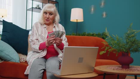 Cheerful-satisfied-elderly-grandmother-freelancer-woman-pressing-button-on-laptop-and-counting-money