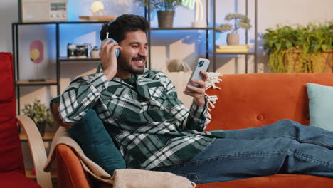 Happy-indian-man-in-wireless-headphones-relaxing-sitting-on-sofa-at-home-listening-favorite-music