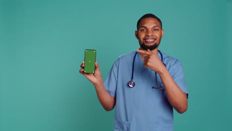 Portrait-of-jolly-male-nurse-pointing-finger-towards-green-screen-smartphone