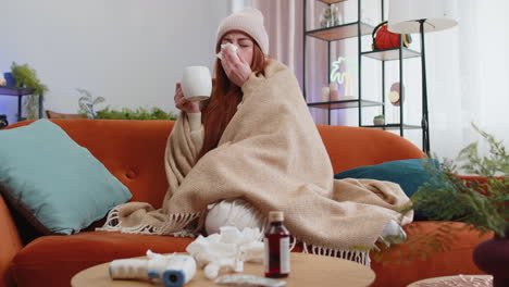 Sick-ill-woman-suffering-from-cold-drinking-hot-tea-sitting-on-couch-sneezes-wipes-snot-into-napkin