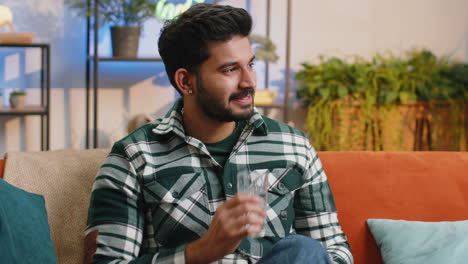 Thirsty-indian-man-sitting-at-home-holding-glass-of-natural-aqua-make-sips-drinking-still-water