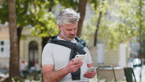 Happy-rich-mature-man-counting-holding-money-dollar-cash-use-smartphone-calculator-app-booking-hotel