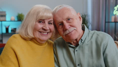 Happy-old-senior-elderly-family-couple-hugging,-laughing,-smiling-looking-at-camera-at-home-sofa