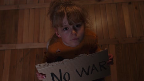 Scared-homeless-toddler-girl-sitting-holding-inscription-No-War,-hiding-from-bombing-attack-at-home