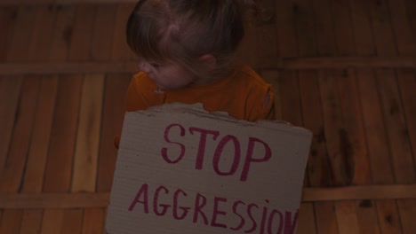 Afraid-homeless-toddler-girl-sitting-holding-inscription-Stop-Aggression,-hiding-from-bombing-attack