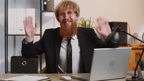 Businessman-working-on-laptop-smiling-friendly-at-camera-and-waving-hands-gesturing-hello-at-office