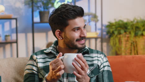 Handsome-smiling-indian-man-drinking-a-cup-of-coffee-or-herbal-tea-sitting-at-home-couch,-break-time