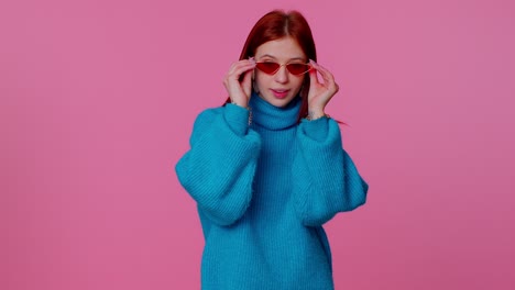 Seductive-cheerful-redhead-girl-in-blue-sweater-wearing-sunglasses,-charming-smile-on-pink-wall