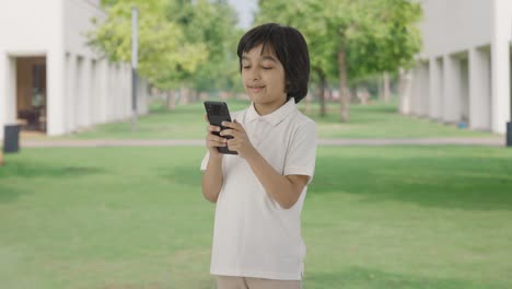 Niño-Indio-Feliz-Enviando-Mensajes-De-Texto-A-Alguien-Por-Teléfono-En-El-Parque