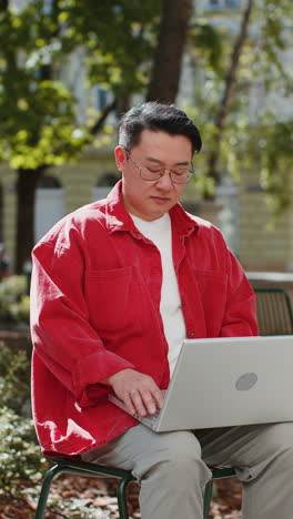 Asian-man-freelancer-working-online-distant-job-with-laptop-browsing-website-sitting-on-city-street