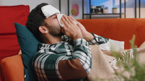 Sick-ill-indian-man-suffering-from-cold-or-allergy-lying-on-home-sofa-sneezes-wipes-snot-into-napkin