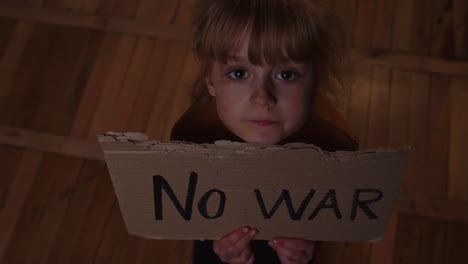 Afraid-homeless-toddler-girl-sitting-holding-inscription-No-War,-hiding-from-bombing-attack-at-home