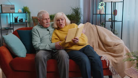 Senior-family-couple-man-woman-enjoying-reading-interesting-book,-talking,-laughing-at-home-on-sofa
