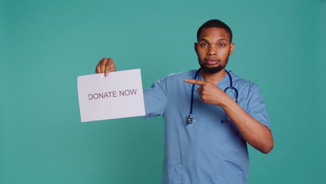 Portrait-of-male-nurse-holding-donate-now-sign,-inviting-people-to-contribute