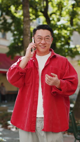 Happy-Asian-man-having-remote-conversation-talking-on-smartphone,-good-news-gossip-in-city-street