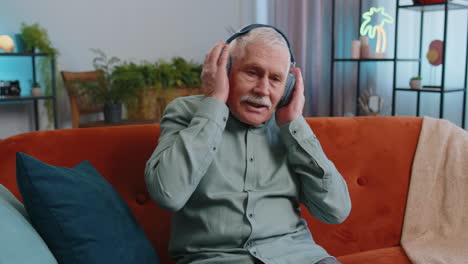 Feliz-Abuelo-Con-Auriculares-Inalámbricos-Relajándose-Se-Sienta-En-El-Sofá-De-Casa-Escuchando-Su-Música-Favorita