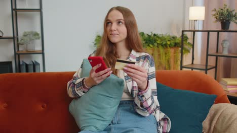 Woman-sitting-at-home-using-credit-bank-card-and-smartphone-while-transferring-money-online-shopping