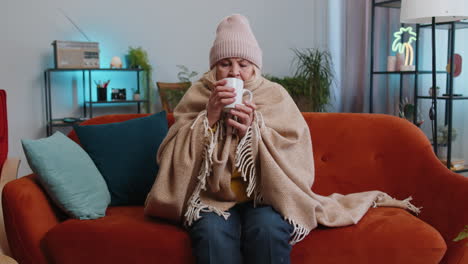 Sick-senior-grandmother-woman-wrapped-in-plaid-sit-alone-shiver-from-cold-on-sofa-drinking-hot-tea