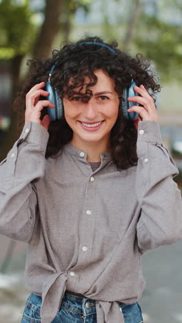 Portrait-of-happy-smiling-woman-listening-music-taking-off-headphones-looking-at-camera-outdoors