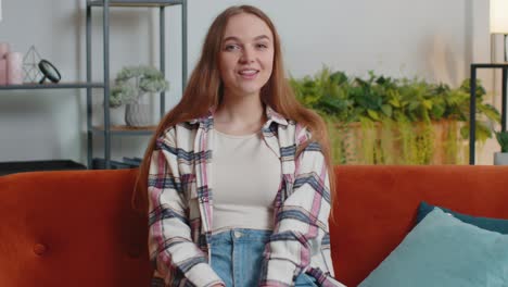 Young-woman-smiling-friendly-at-camera,-waving-hands-gesturing-hello,-hi,-greeting-at-home-on-sofa