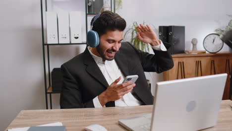 Indian-business-man-in-headphones-listening-favorite-disco-music,-working-on-laptop-at-home-office