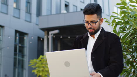 Glücklicher-Indischer-Geschäftsmann-Tippt-Ferngesteuert-Auf-Einem-Kabellosen-Laptop-Computer-Und-Surft-Im-Freien-Im-Internet