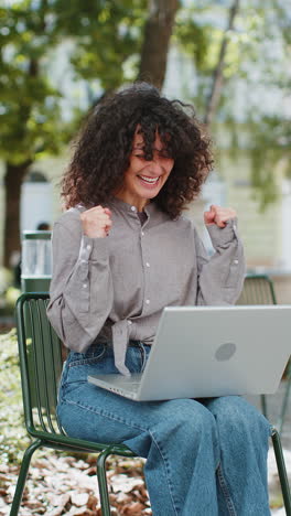 Caucasian-happy-woman-working-on-laptop-celebrate-success-win-money-sitting-on-urban-street-in-city