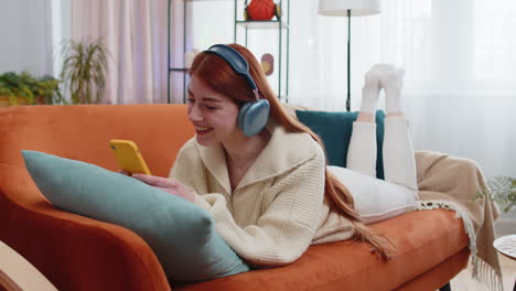 Mujer-Joven-Feliz-Con-Auriculares-Inalámbricos-Relajándose-Tumbada-En-El-Sofá-De-Casa-Escuchando-Su-Música-Favorita