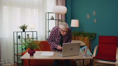 Senior-grandfather-man-working-on-laptop-computer-making-notes-on-sheet-of-paper-calculating-bills