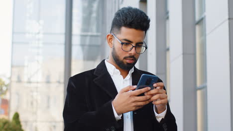 Smiling-business-man-using-smartphone-typing-text-answering-messages-chatting-online-in-city-street