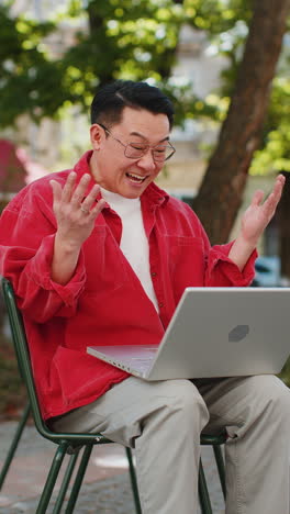 Asian-happy-man-working-on-laptop-celebrate-success-win-money-sitting-on-urban-street-in-city