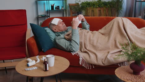 Sick-ill-elderly-man-suffering-from-cold,-allergy-lying-on-home-sofa-sneezes-wipes-snot-into-napkin