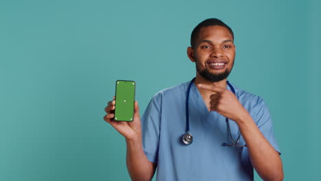 Retrato-De-Un-Alegre-Enfermero-Apuntando-Con-El-Dedo-Hacia-Un-Teléfono-Inteligente-De-Pantalla-Verde