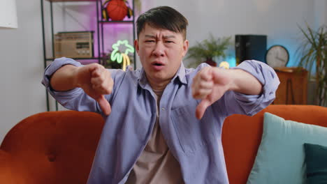 Upset-asian-man-showing-thumbs-down,-dislike-bad-work,-disapproval,-dissatisfied-feedback-at-home