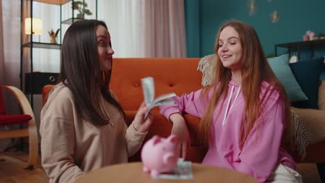 Girls-friends-siblings-sitting-on-floor-and-take-turns-dropping-dollar-banknote-into-piggy-bank