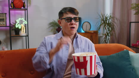 Asian-man-sits-on-sofa-eating-popcorn-and-watching-interesting-TV-serial,-sport-game-online-at-home