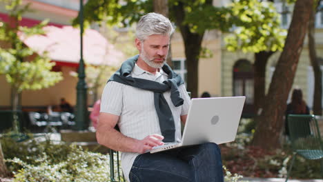 Middle-aged-man-opens-laptop-start-working-sends-messages-sitting-on-chair-in-urban-city-street