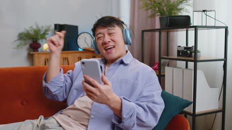 Happy-asian-man-in-wireless-headphones-relaxing-sitting-on-sofa-at-home-listening-favorite-music