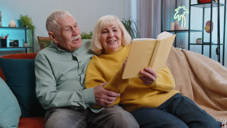 Senior-family-couple-man-woman-enjoying-reading-interesting-book,-talking,-laughing-at-home-on-sofa