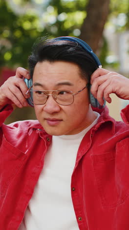 Portrait-of-happy-smiling-Asian-man-listening-music-taking-off-headphones-looking-at-camera-outdoors