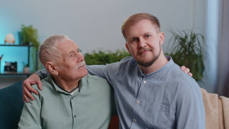 Portrait-of-mature-grandfather-with-young-adult-man-grandson-smiling-happy-embracing-hugging-at-home